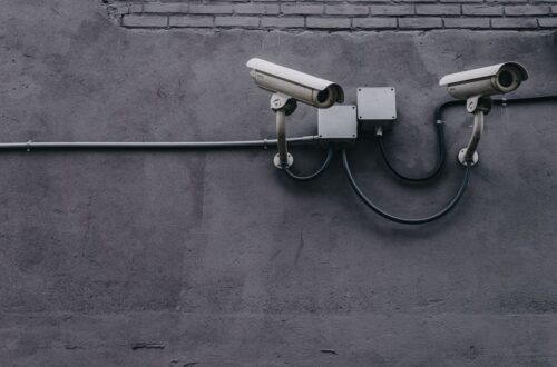 Two white security cameras mounted on a grey exterior wall with conduit boxes